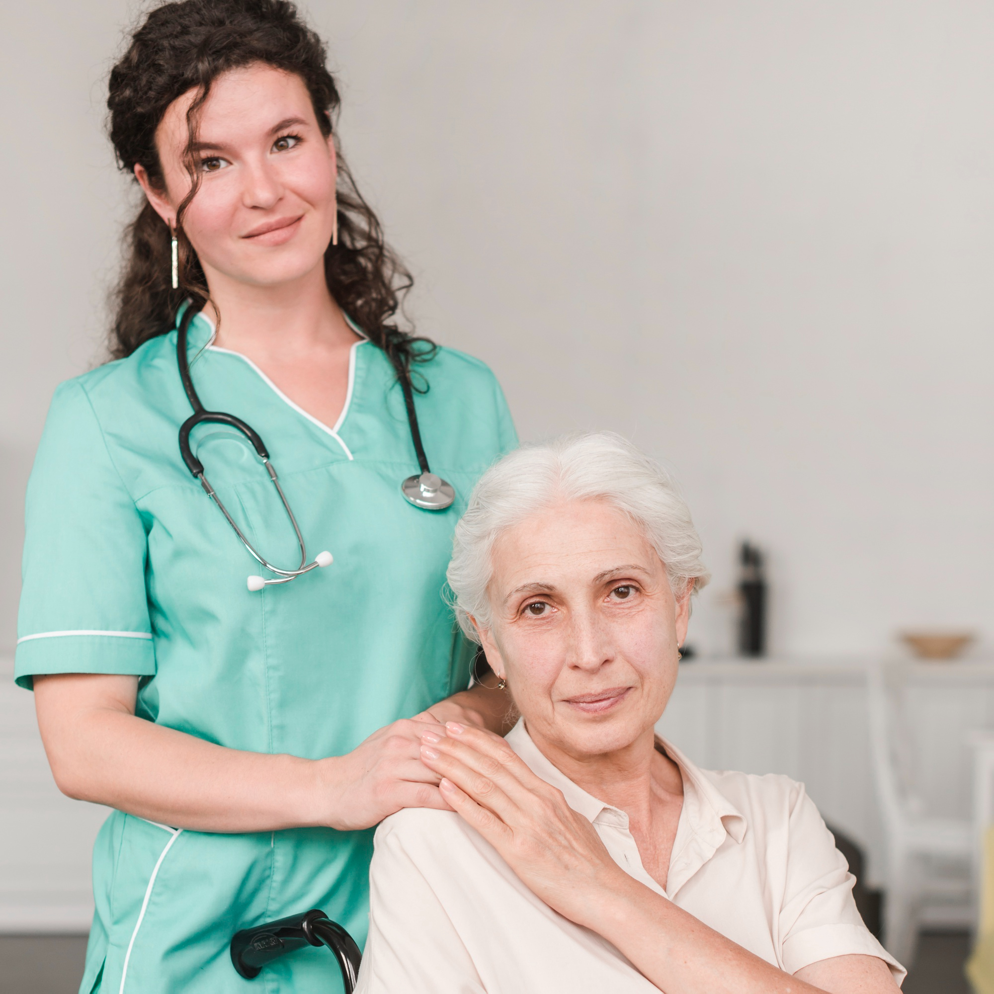 Smiling female carer in green scrubs standing beside an elderly woman seated at home, gently resting her hands on the client’s shoulders in a supportive pose.
