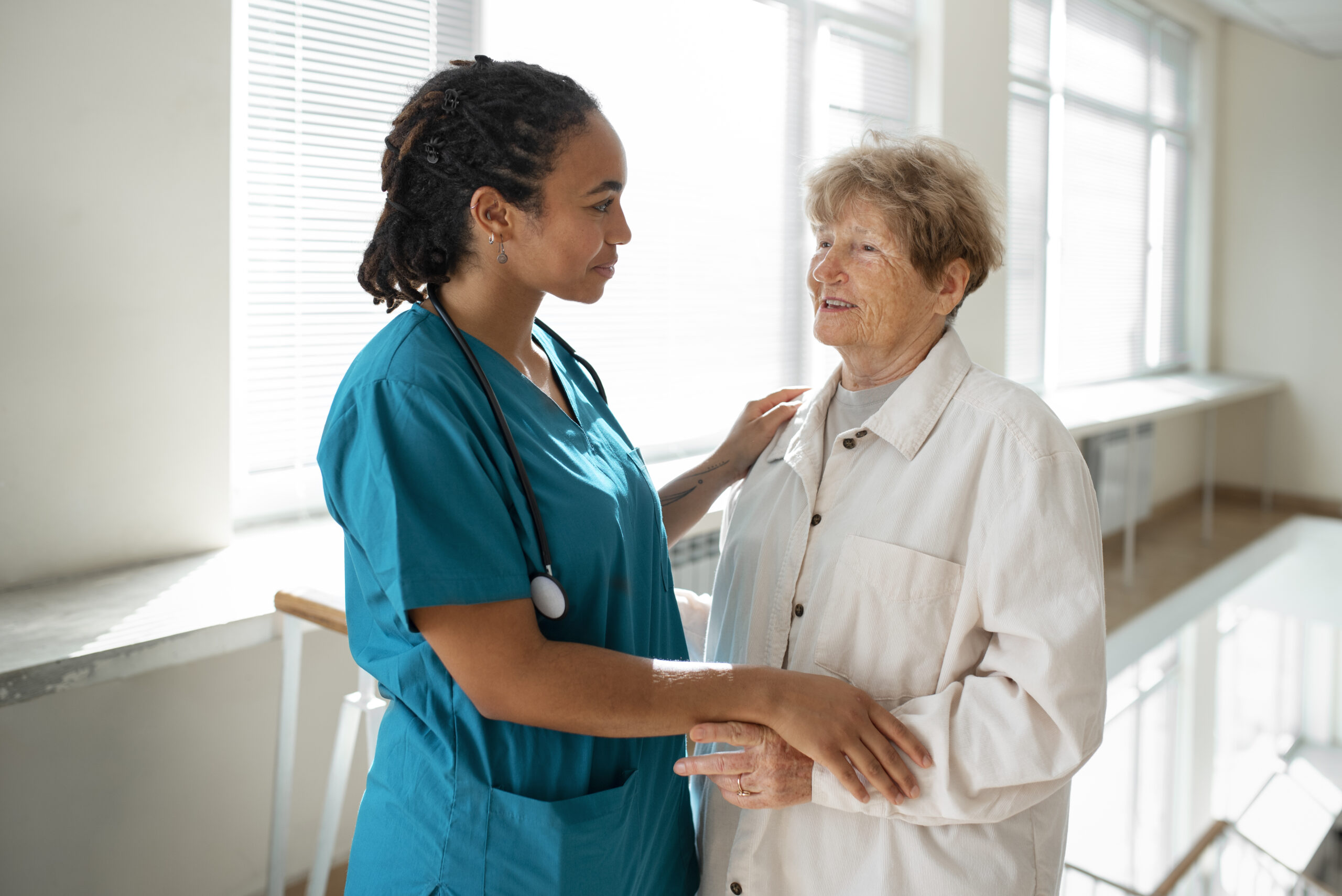 Professional carer in teal scrubs gently holding hands with an elderly woman in her home, offering reassurance and support.